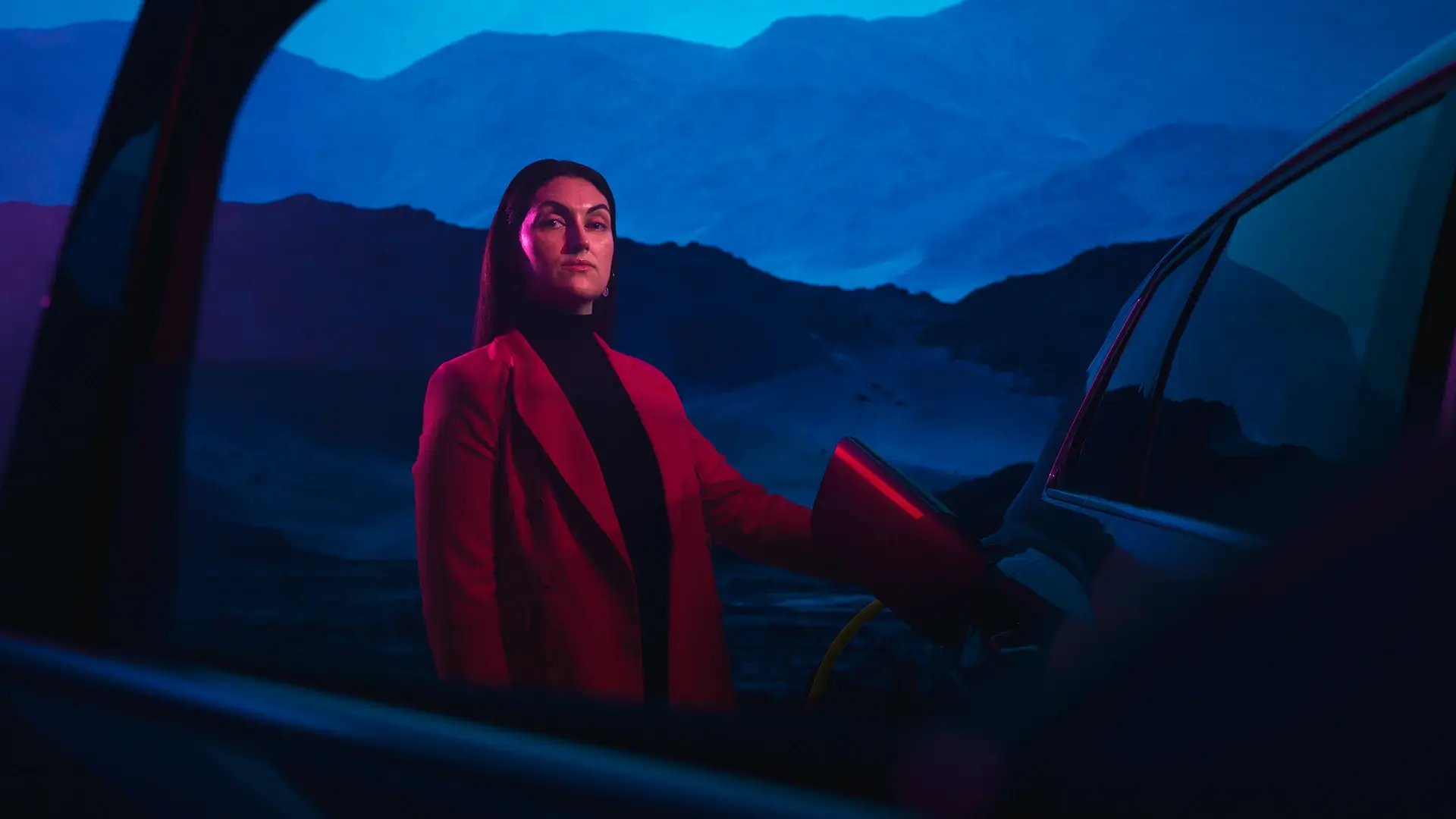 Frau im Business-Outfit lädt E-Auto vor blauen Nachthimmel und Bergkulisse