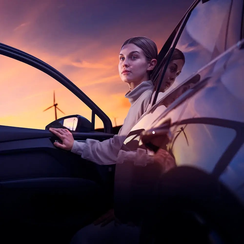 Självförtroendefull kvinna i elbil med vindkraftverk i soluppgången