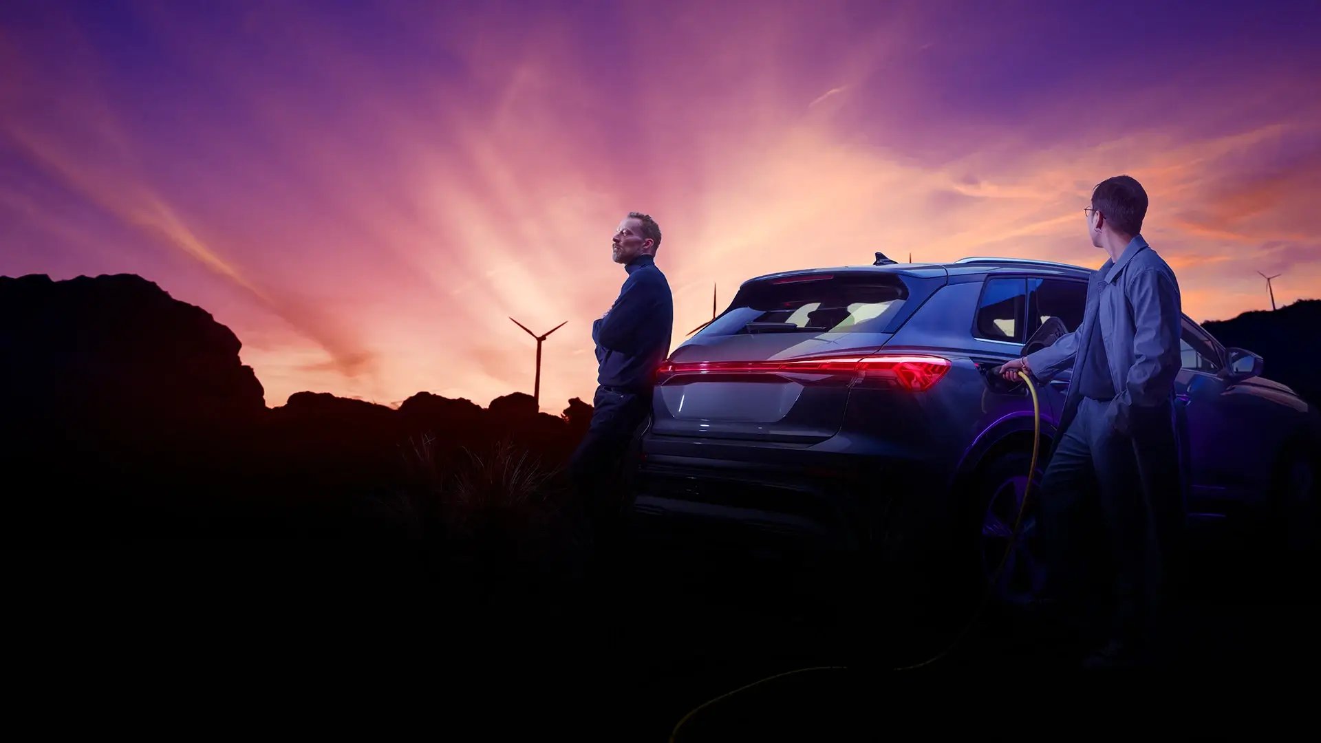 Sonnenuntergang und Windrad und im Vordergrund zwei Männer die ein elektrisches SUV E-Auto laden