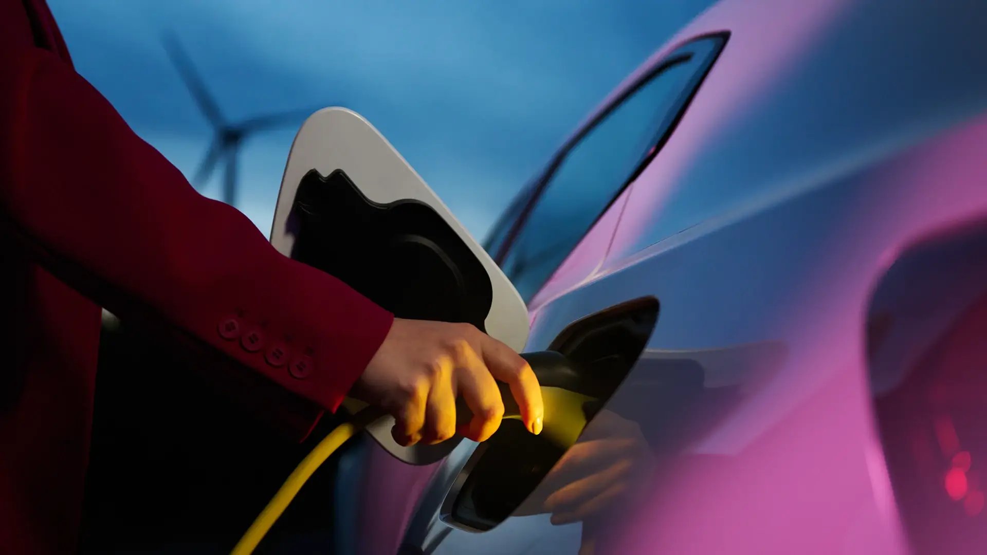 Hand von Frau steckt Ladekabel an E-Auto mit Windkraftanlage im Hintergrund