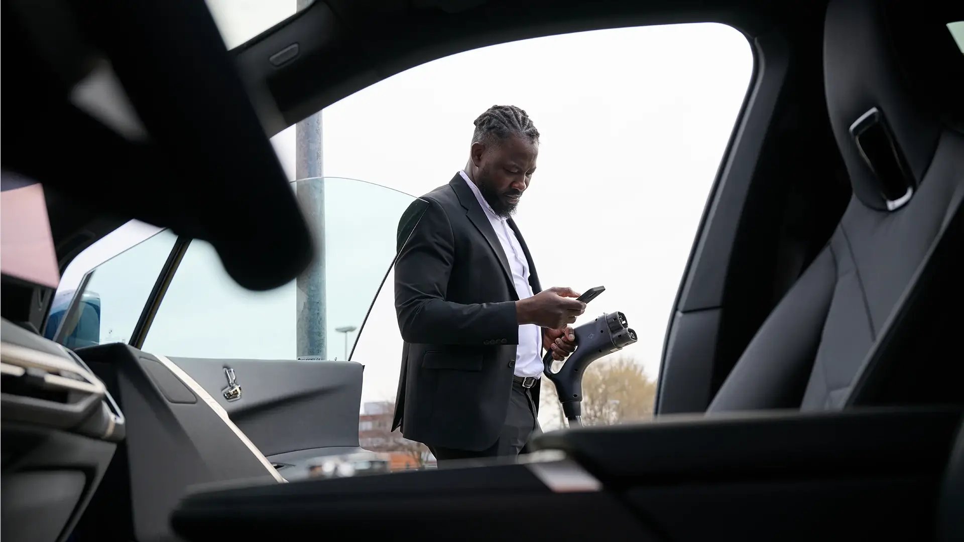 Business Mann benutzt Smartphone neben dem E-Auto und hält CCS Ladekabel in seiner linken Hand