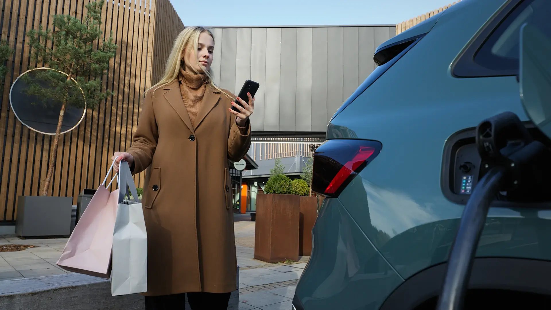 Frau mit Einkaufstüten benutzt Handy um E-Auto zu laden