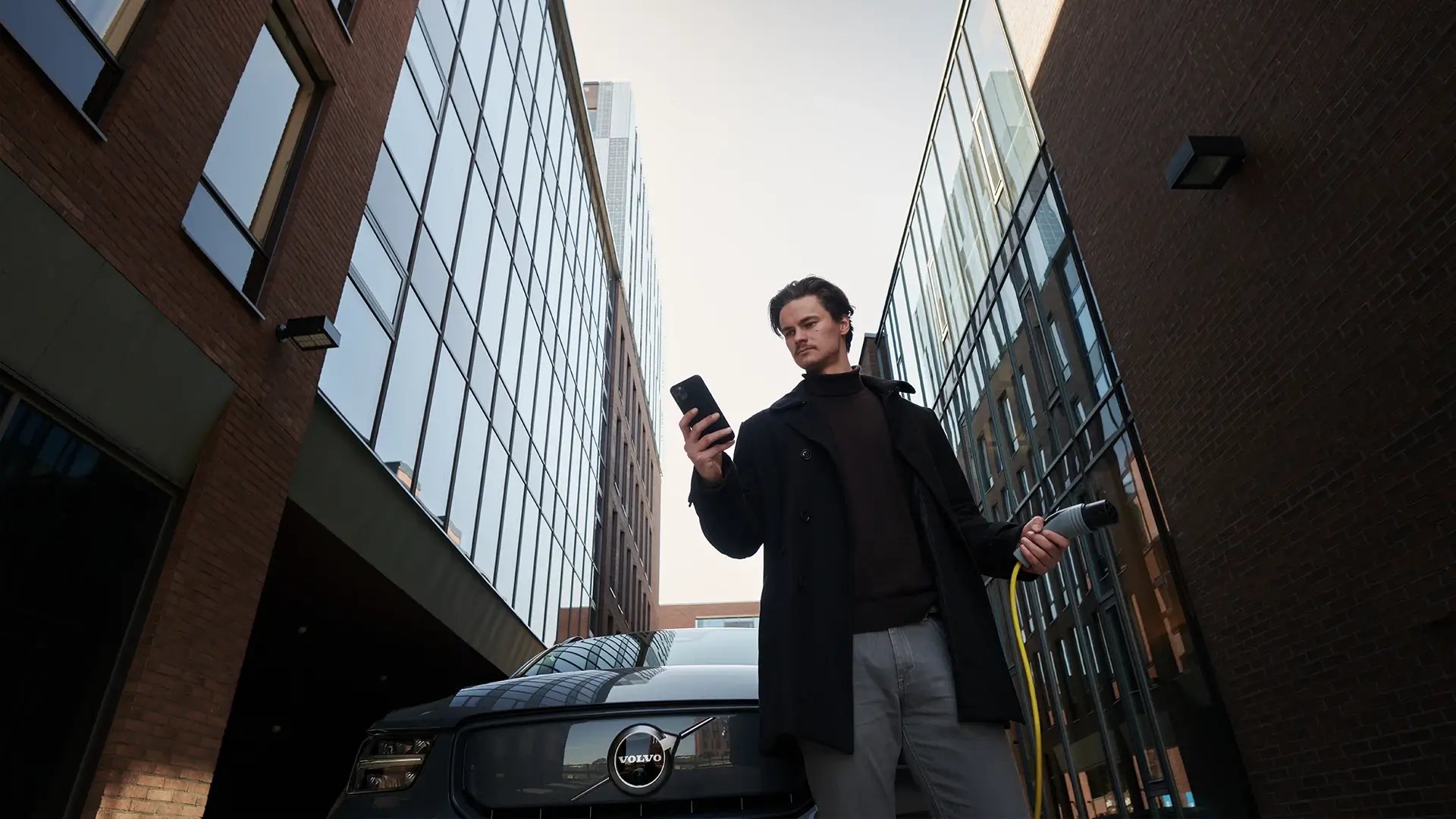 Mann startet Ladevorgang für sein E-Autos über eine App in seinem Smartphone und hält dabei gelbes Ladekabel in der Hand
