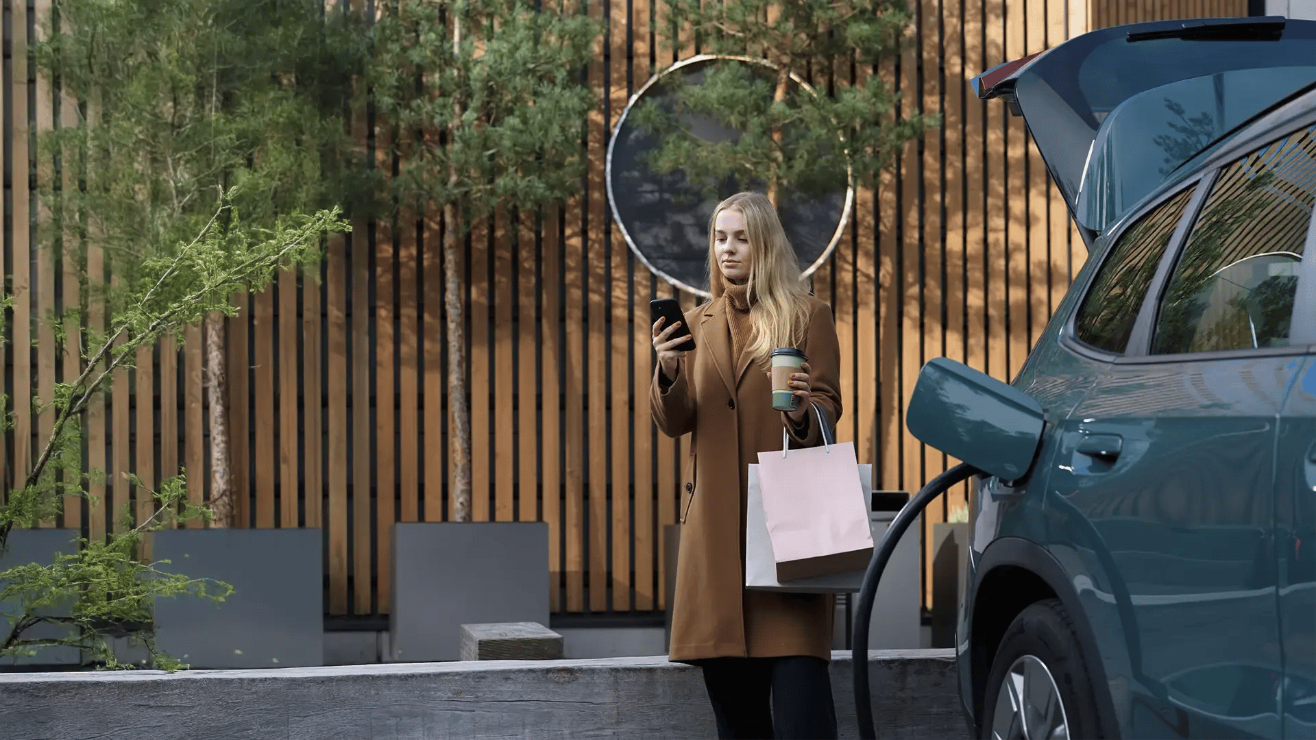A woman standing beside an electric vehicle while it charges, holding a coffee cup and shopping bags