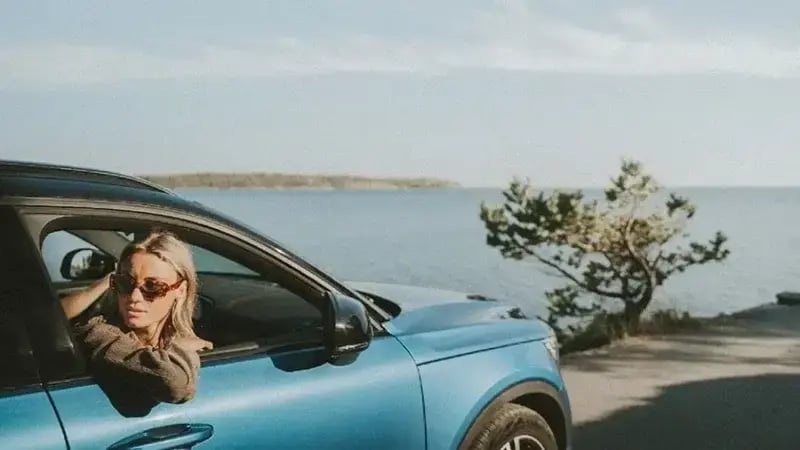 Woman wearing sunglasses sitting in a blue car by a coastal road with sea in the background