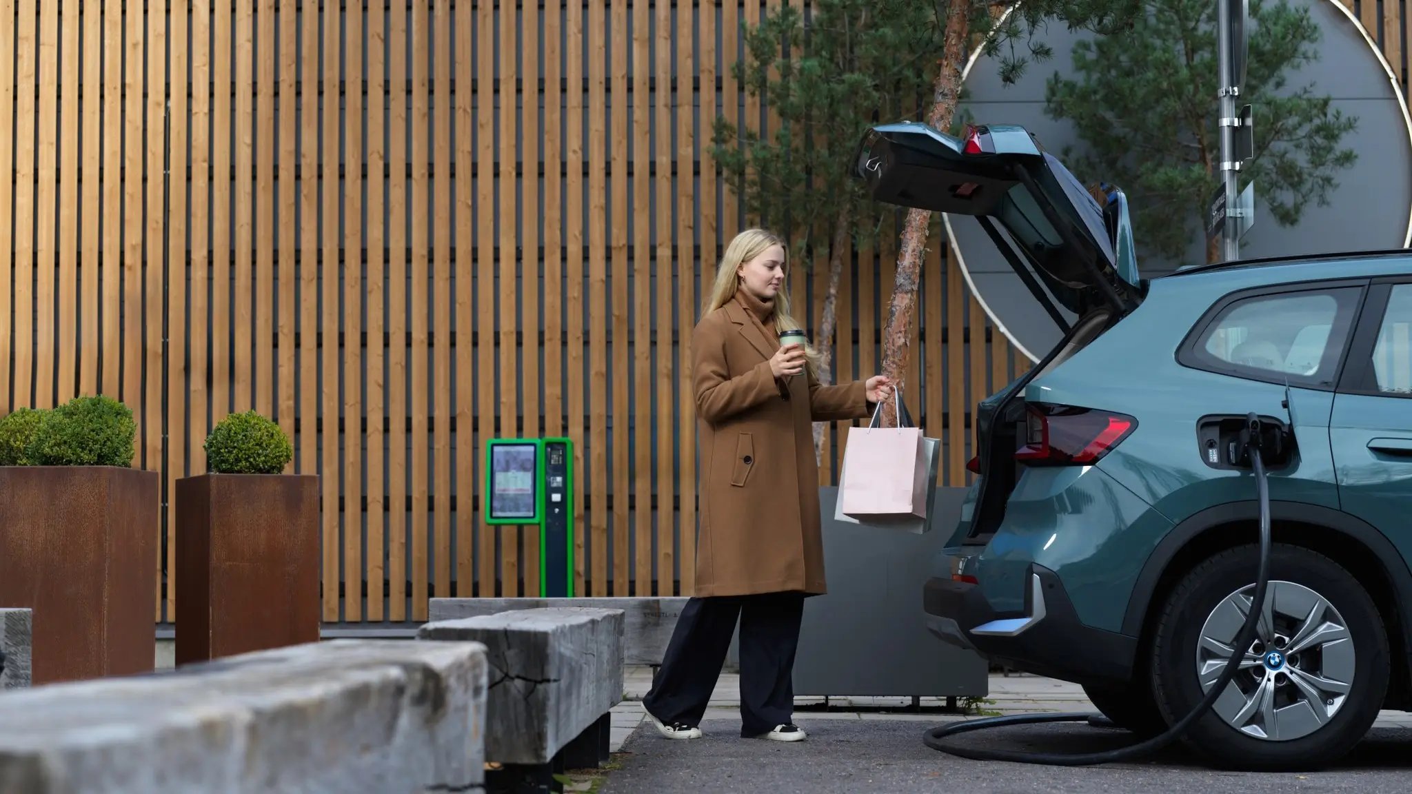 Junge Frau legt Einkaufstüten in den Kofferraum ihres E-Autos, das gerade an einer Ladestation lädt