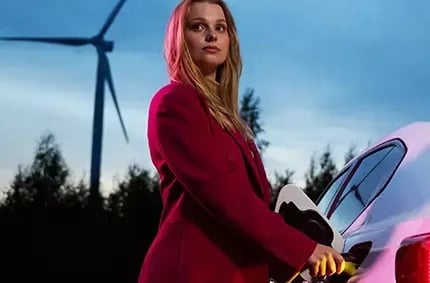 Foto på kvinna, hållandes på en laddkontakt som är ansluten till en elbil, med skog och ett vindkraftverk i bakgrunden