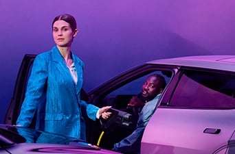 Foto på kvinna med en laddkontakt i ena handen, ståendes bredvid en elbil i vilken en man sitter med en öppen dörr