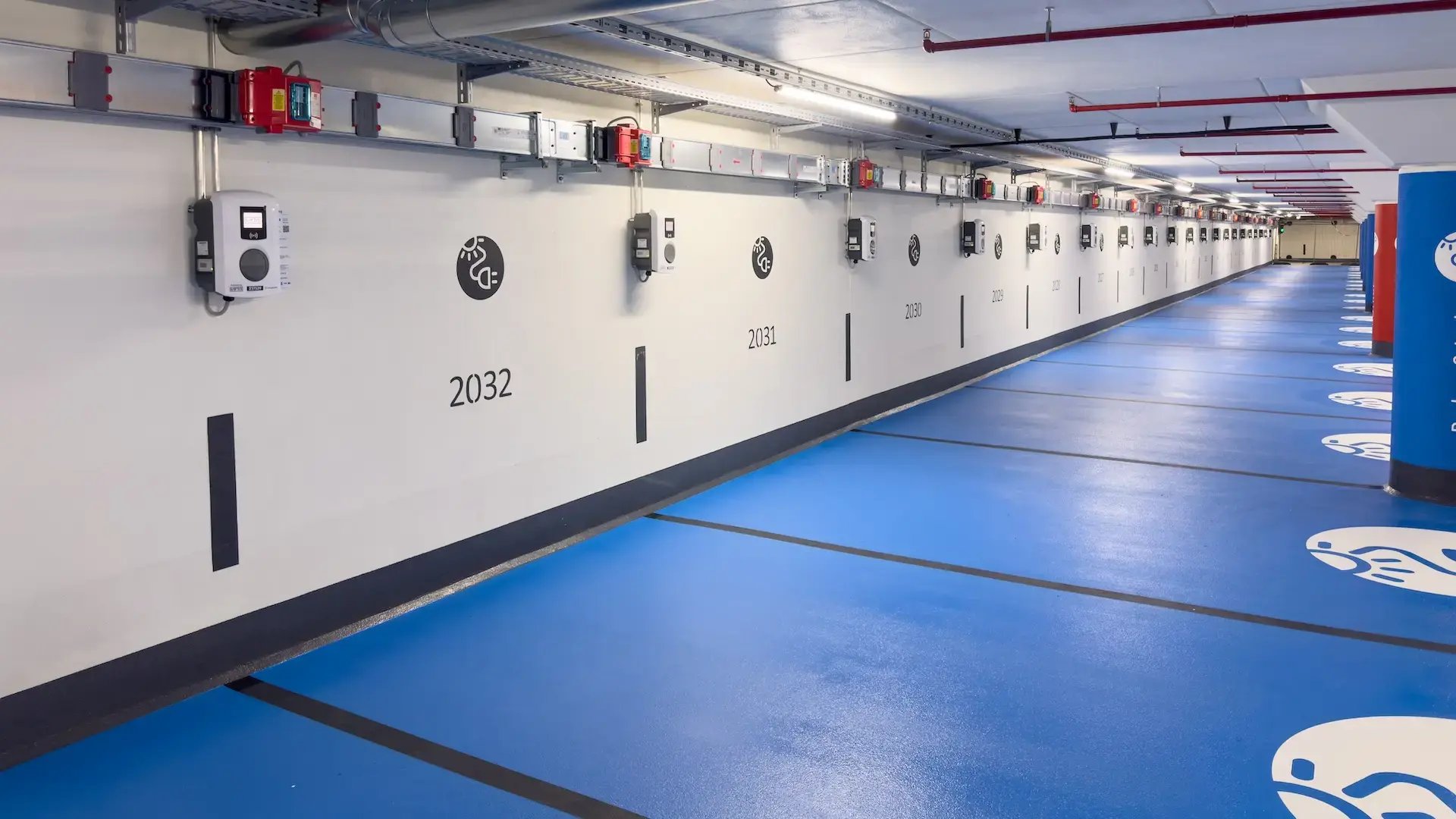 Modernes unterirdisches Parkhaus mit blau markierten Parkplätzen, jeder mit einer Elektroauto-Ladestation an der Wand ausgestattet. Der Standort ist das Parkhaus Rathaus in Mainz, betrieben von PMG Parken in Mainz GmbH.