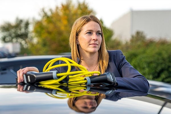 Foto på Aurélie Allier, Direktör för Clos du Chêne-handelsområdet, stående bredvid en elbil, med en laddkabel på taket av bilen