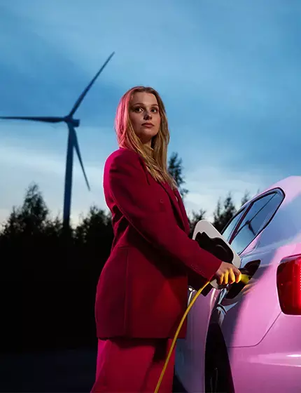 Bild på elbil och kvinna som står med en laddkabel ansluten till bilen och i bakgrunden ser man en skog och ett vindkraftverk