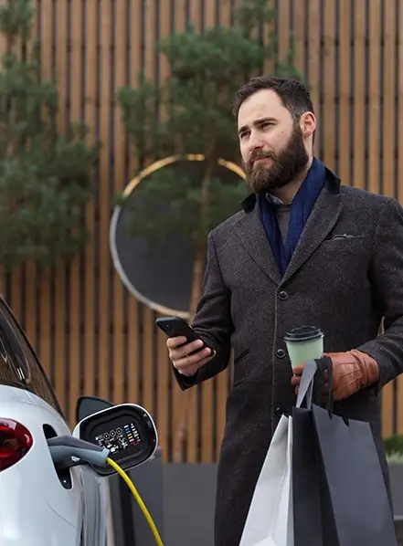 Foto på en man med shoppingpåsar ena handen och en mobiltelefon i andrahanden, ståendes bredvid en elbil med en laddkabel ansluten till den