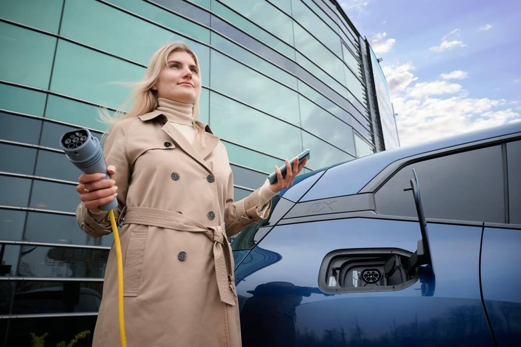 Foto på kvinna som håller en mobiltelefon i ena handen och en laddkabel till en elbil i den andra