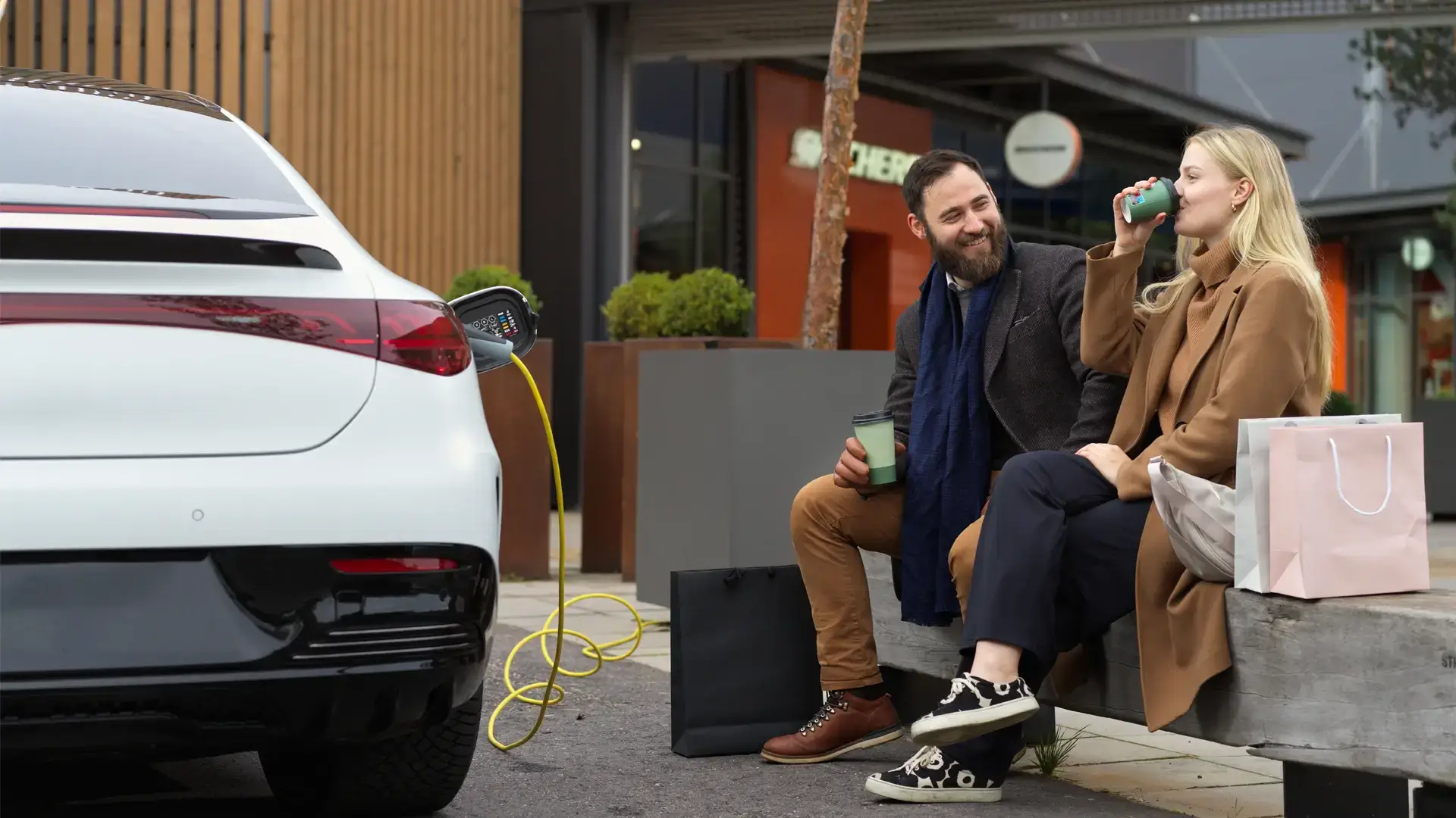 Nainen ja mies isutuvat puistonpenkillä juomassa kahvia samalla kun lataavat sähköautoa