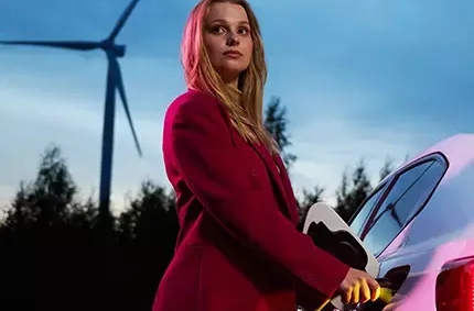 Bild på kvinna som har anslutit en elbilsladdarkontakt till en elbil, med en skog och ett vindkraftverk i bakgrunden
